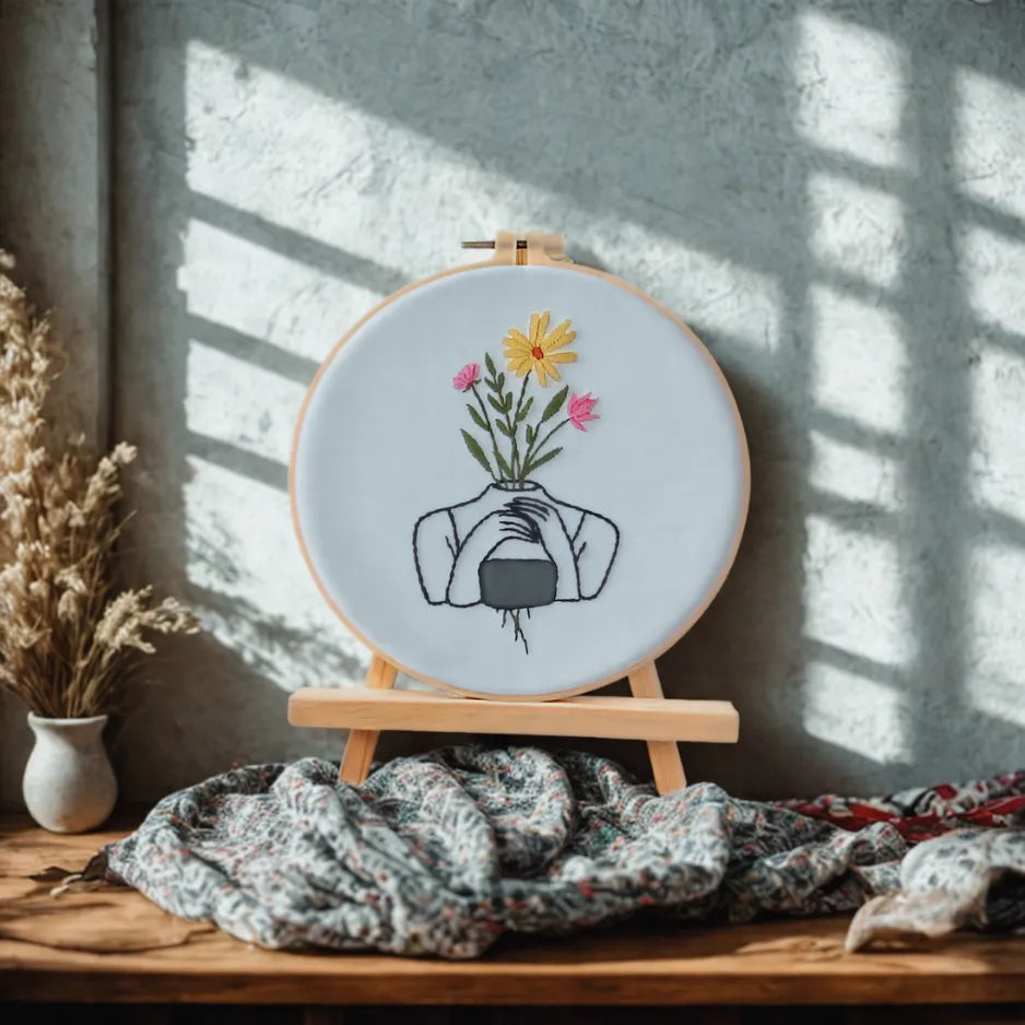 Embroidery hoop with floral design on a wooden stand against a textured wall.