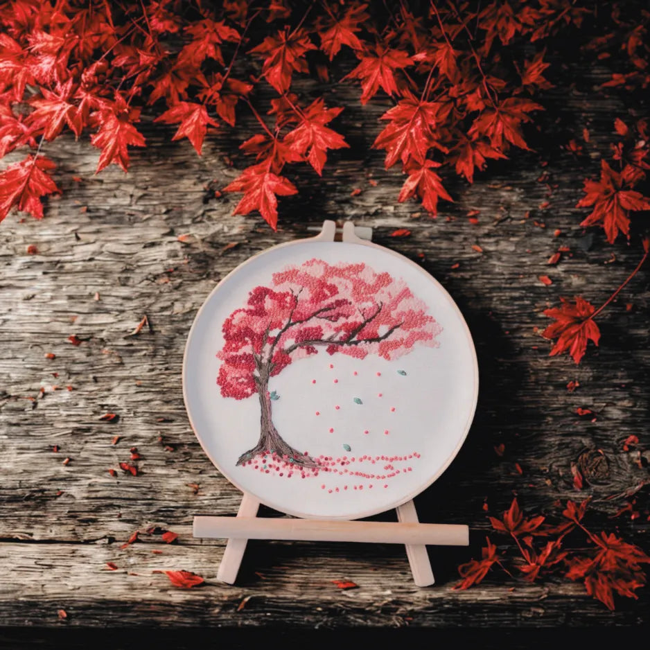Embroidery hoop with cherry blossom tree design on a wooden surface with red leaves.