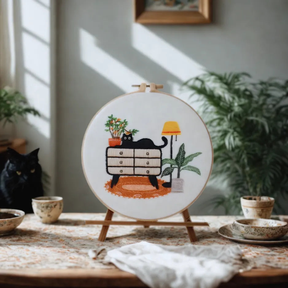 Embroidery hoop with a cat design on a wooden table with plants and bowls.