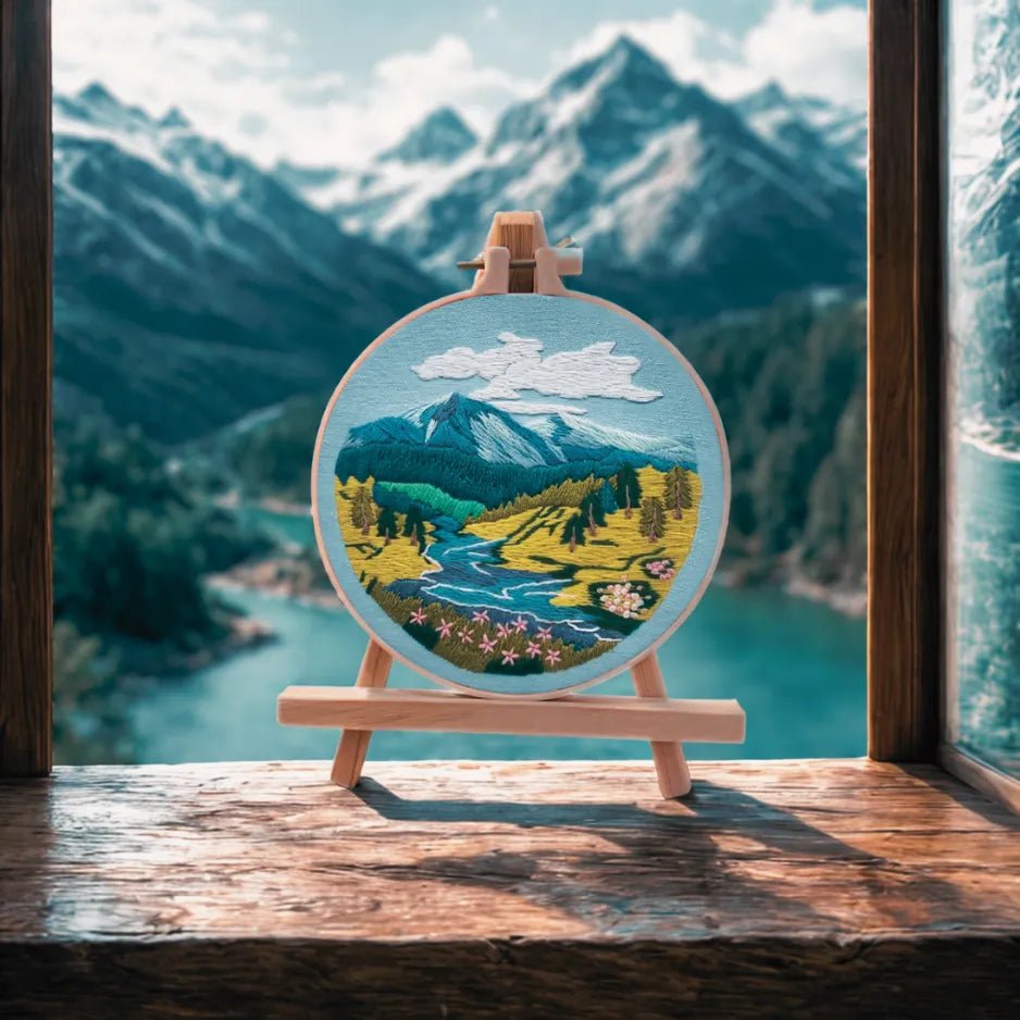 Embroidery hoop with a mountain landscape design in front of a window with a mountain view.