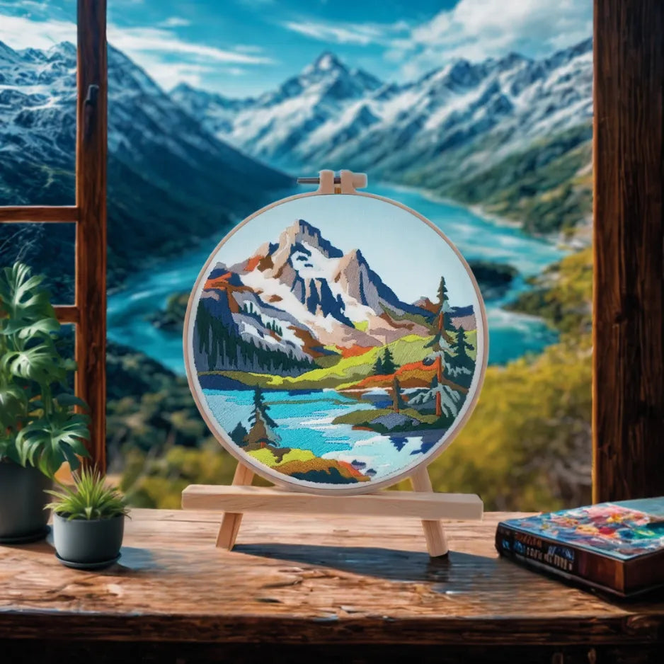 Embroidery hoop with mountain landscape design in front of a window with a view of mountains and a lake.