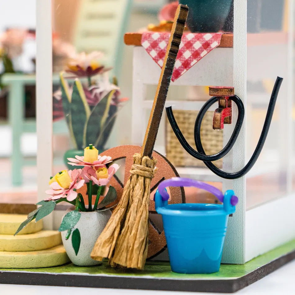 Miniature broom and blue bucket on a table with flowers and a checkered cloth in the background.