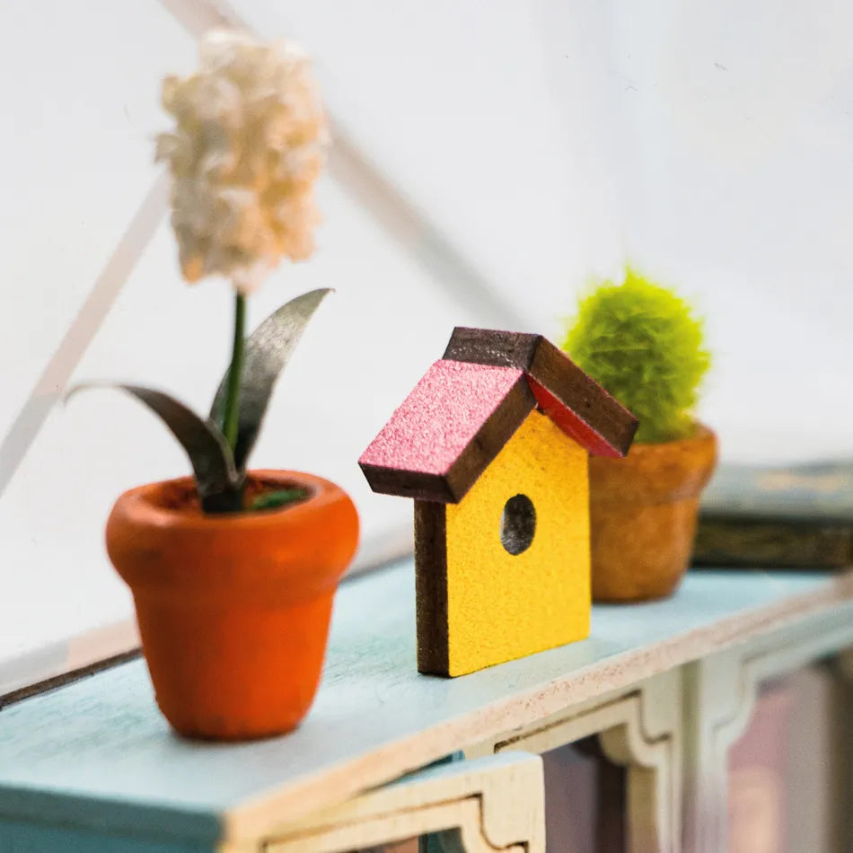 Small wooden birdhouse with pink roof and yellow body on a surface with potted plants.