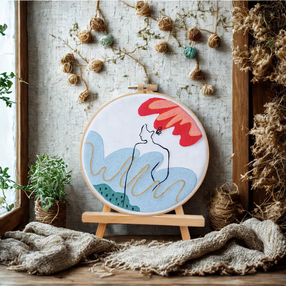 Embroidery hoop with a colorful design of a woman with a red hat on a wooden stand, surrounded by natural elements.