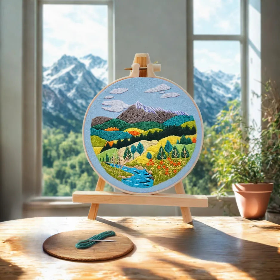 Embroidery hoop with a mountain landscape design on a wooden easel in front of a window with a mountain view.