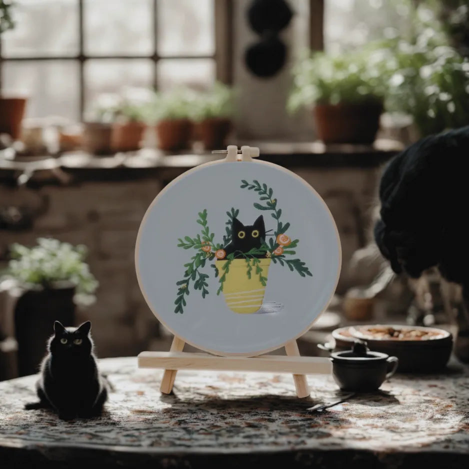Embroidery hoop with a black cat design on a table in a cozy room with plants.
