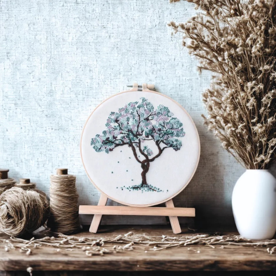 Embroidery hoop with a tree design on a wooden stand against a light blue wall.