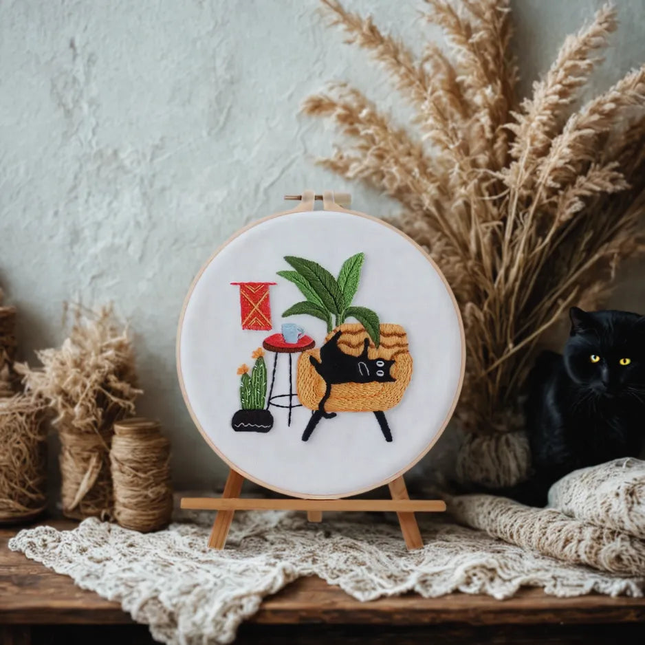 Embroidery hoop with a black cat design on a wooden surface with decorative elements.